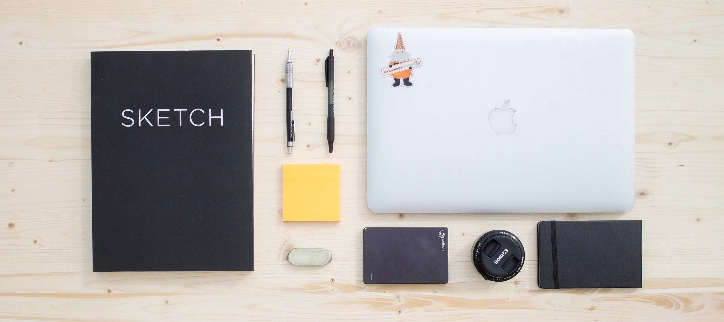 flatlay of sketchbook, small notepad, external hardrive, Macbook, Canon camera lens, drafting pencil, ballpoint pen, eraser, and sticky notes